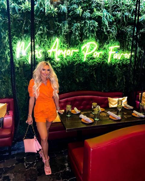Blonde girl in an orange romper on a greenery wall with a neon sign.